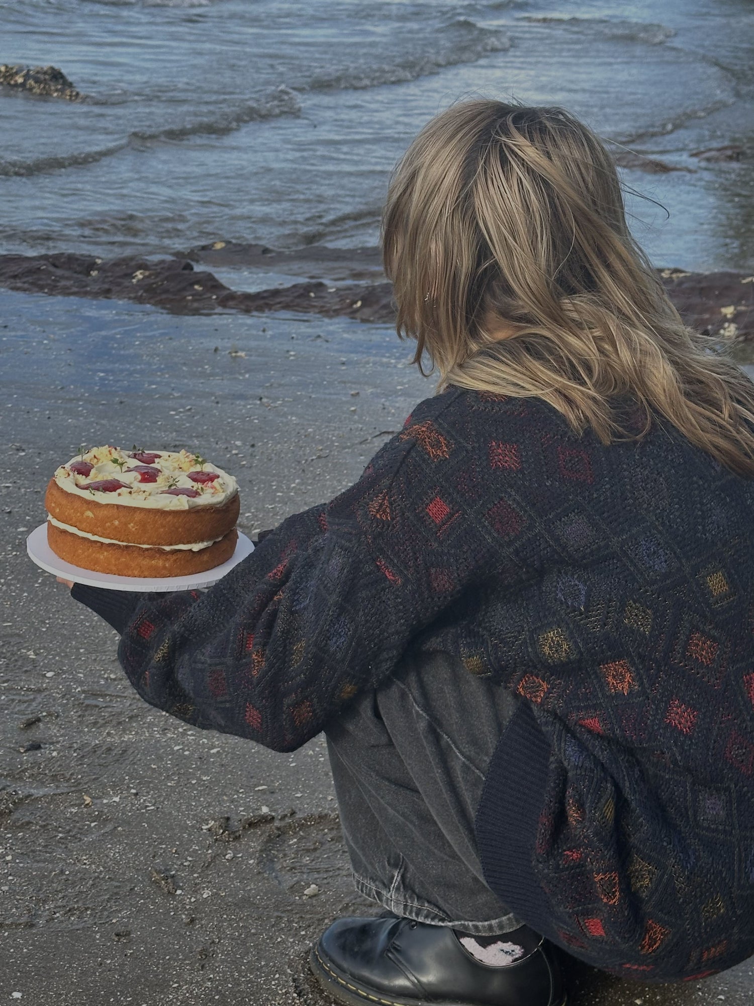 Paloma from Zi Sweet holding a strawberry and mascarpone sponge cake and looking out to sea at the beach.