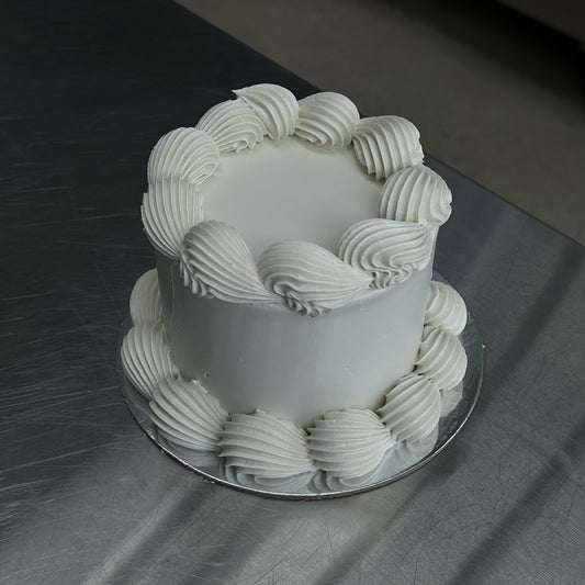 A small mini vintage style sponge cake coated with a white swiss buttercream meringue. The base and top edges are decorated in white with a piped shell.