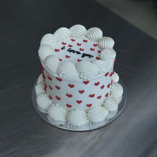 A small mini vintage style sponge cake coated with a white swiss buttercream meringue. The base and top edges are decorated in white with a piped shell and the sides and top are decorated with lots of mini red piped hearts.  There is black writing on the top of the cake saying "I love you".
