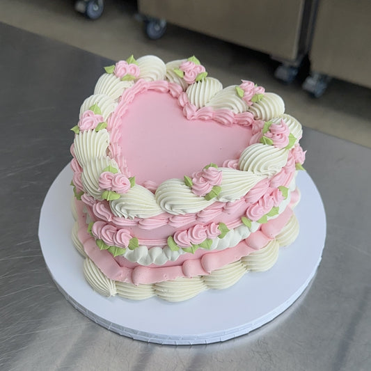 A large heart shaped vintage style sponge cake coated with a baby pink swiss buttercream meringue. The base and top edges are decorated in white with a piped shell and the sides are intricately decorated with buttercream piping of small flowers.