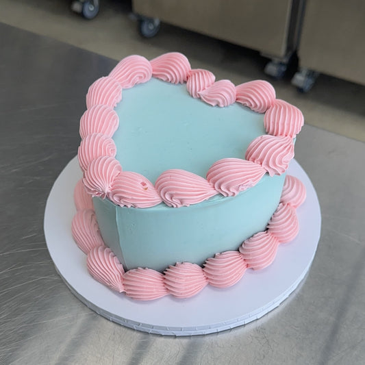 A large heart shaped vintage style sponge cake coated with a pale blue swiss buttercream meringue. The base and top edges are decorated in baby pink with a piped shell.