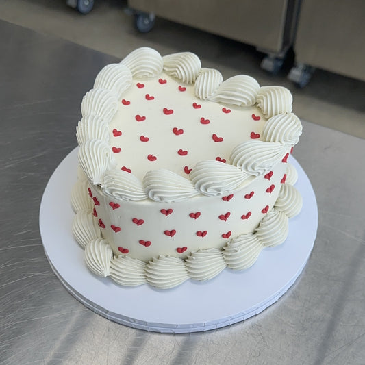 A large heart shaped vintage style sponge cake coated with a white swiss buttercream meringue. The base and top edges are decorated in white with a piped shell and the sides and top are decorated with lots of mini red piped hearts.  