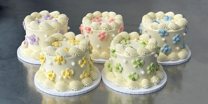 Five small mini cakes all coated in a white Swiss meringue buttercream and decorated with white piped shells around the top and bottom of the cake.  Each cake is decorated with either purple, pink, blue, yellow or green pastel-coloured daisies on the top and sides of the cake.