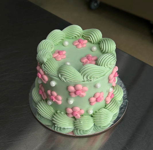 A mini round shaped vintage style sponge cake coated with a light green swiss buttercream meringue. The base and top edges are decorated in green with a piped shell and the sides and top are decorated with lots of pink buttercream daisies and white sugar pearls.