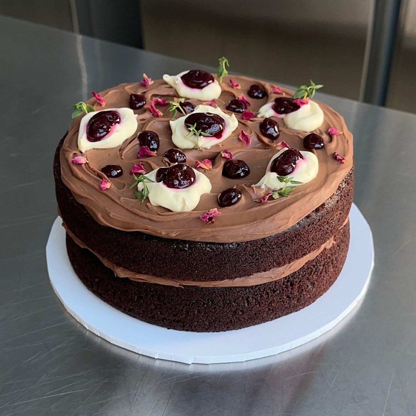 A two-layer chocolate sponge cake with a chocolate ganache filling and topped with a chocolate ganache and dollops of cream, boysenberry jam and a sprinkling of rose petals and thyme to decorate.