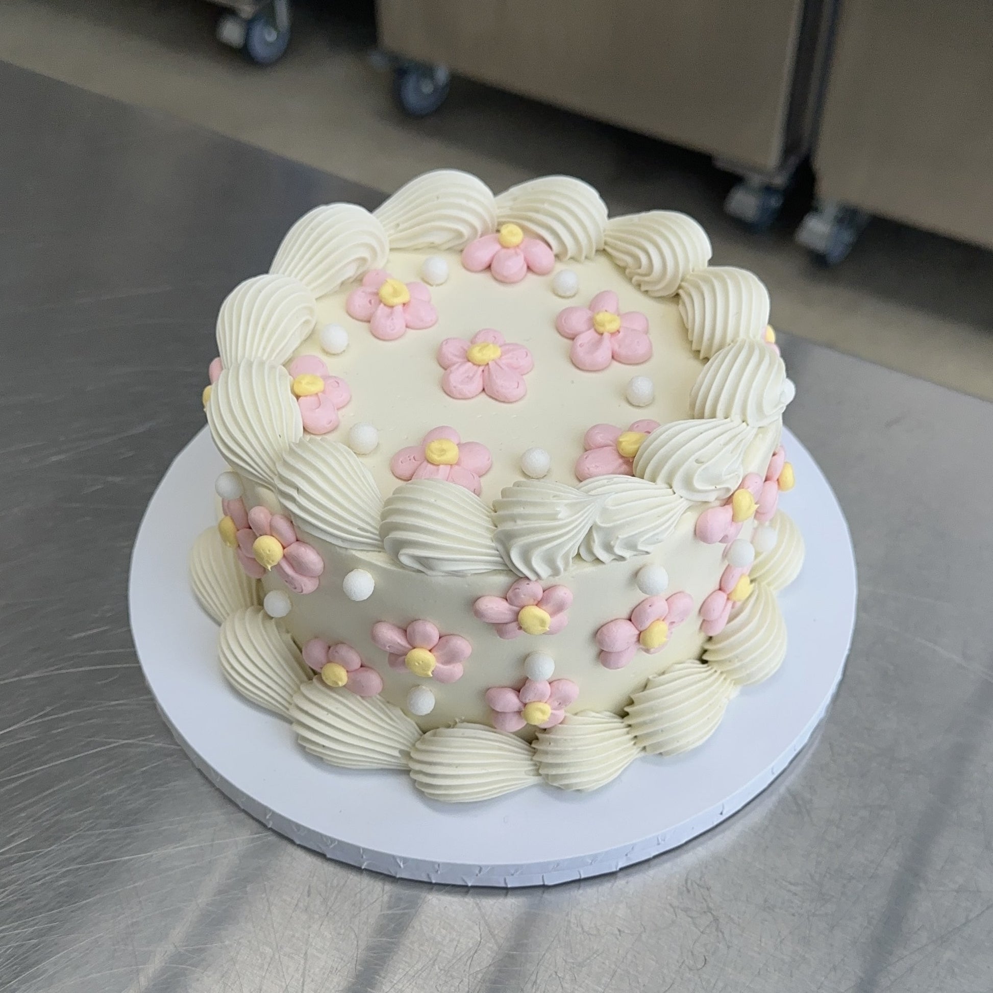 A round shaped vintage style sponge cake coated with a white swiss buttercream meringue. The base and top edges are decorated in white with a piped shell and the sides and top are decorated with lots of pink and yellow buttercream daisies.